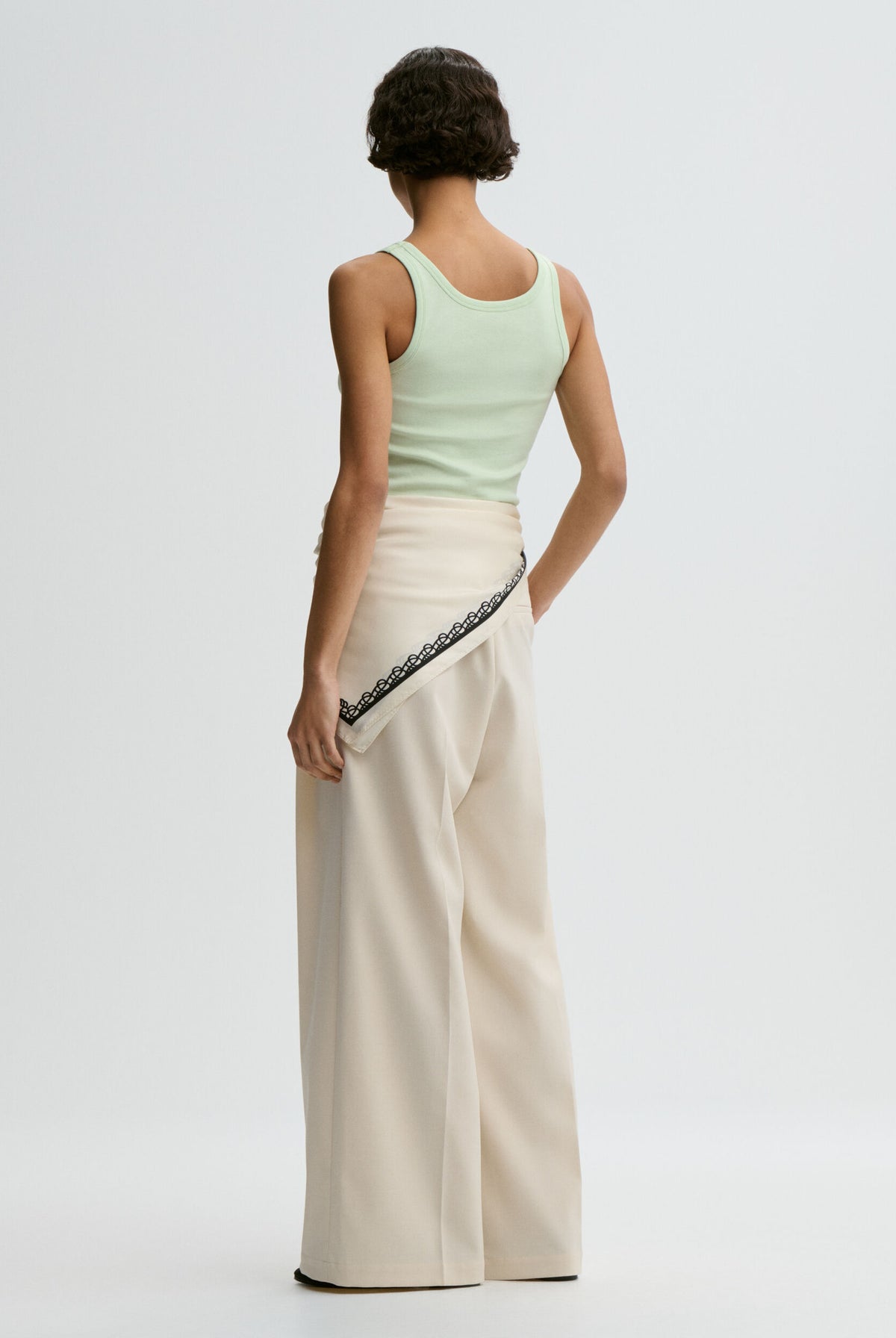 Woman wearing a light green sleeveless top and beige pants with lace trim on a white background