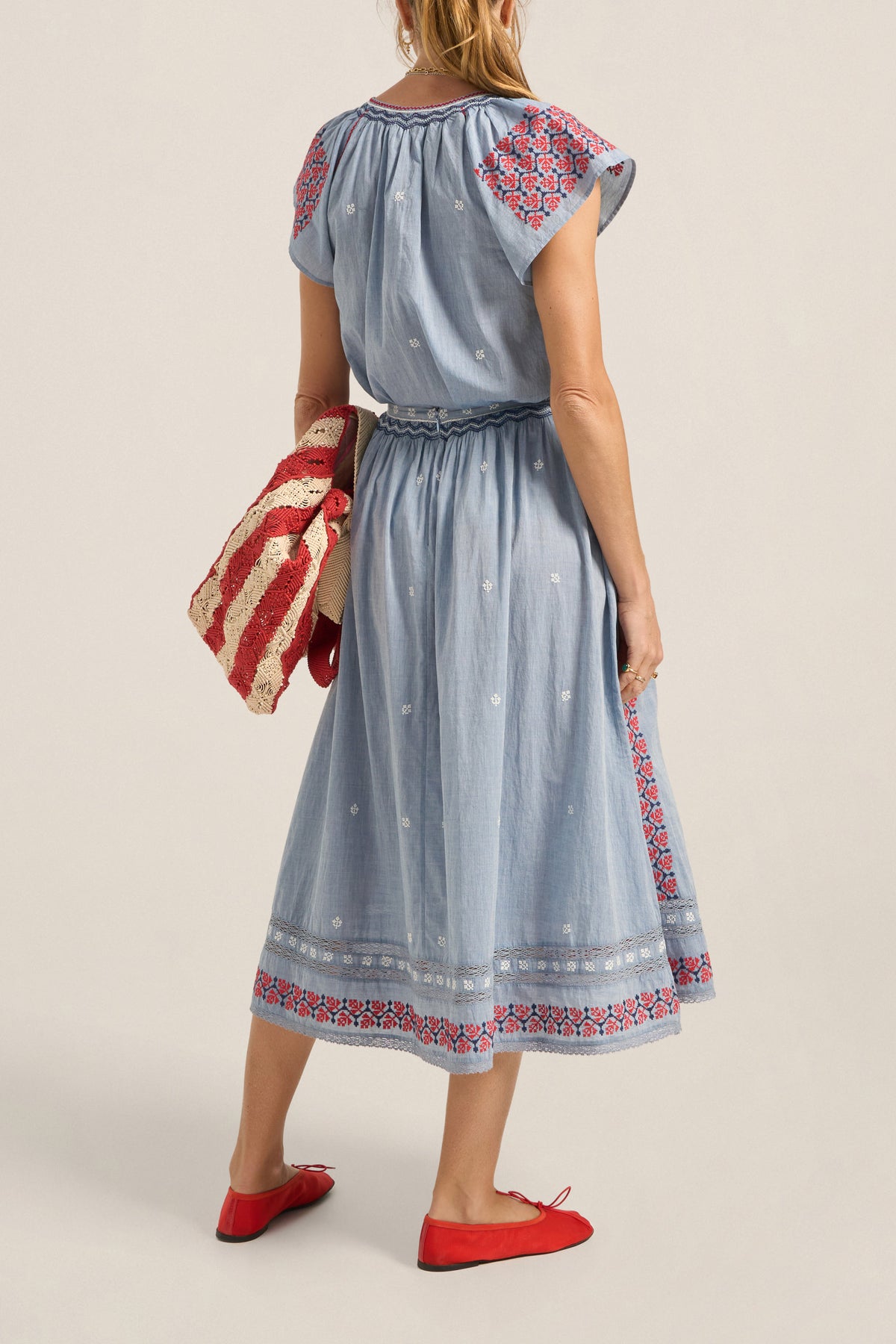 Woman wearing a blue dress with red and white patterned sleeves and hem, holding a red and white bag.