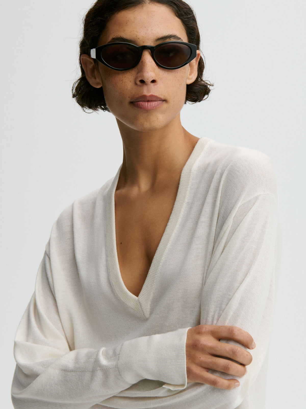 Woman wearing a white sweater and dark sunglasses against a light background