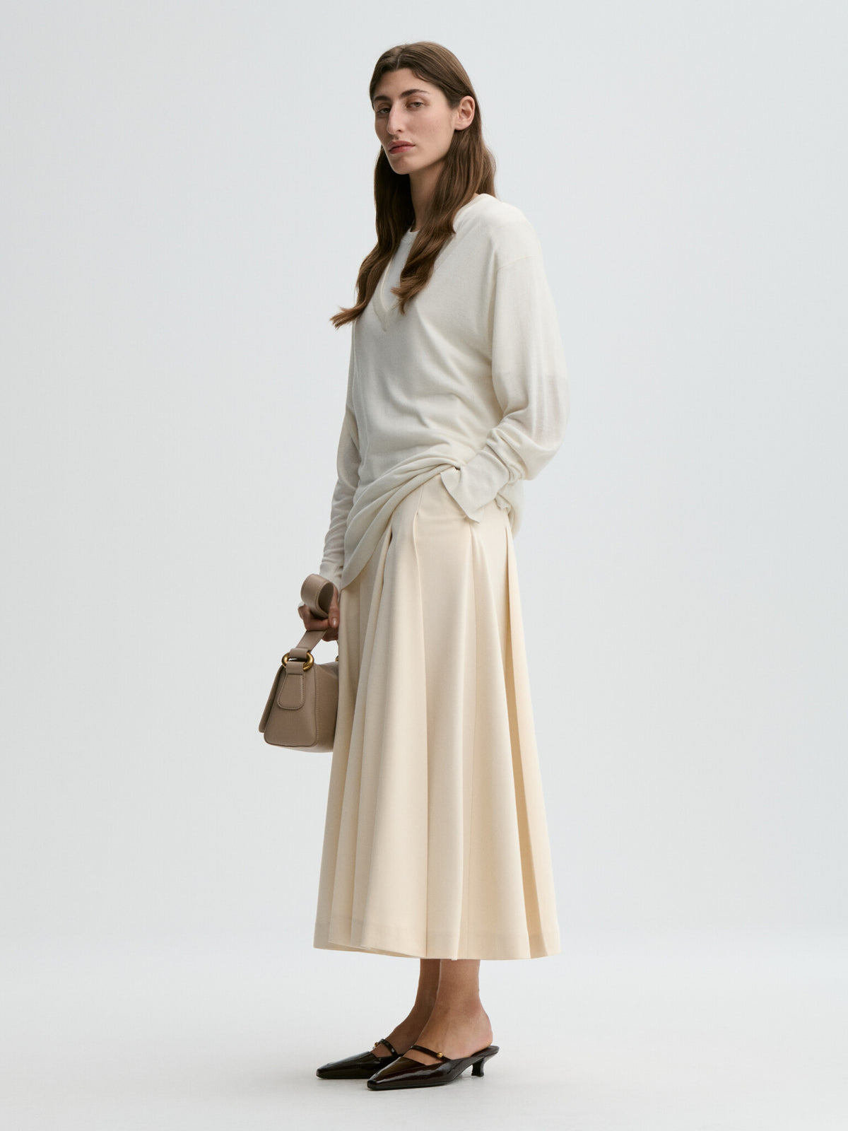 Woman wearing a white sweater and beige skirt holding a beige handbag on a light gray background