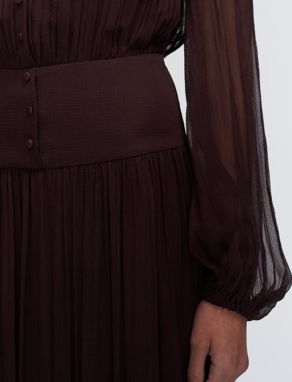 Close-up of a dark brown long-sleeve dress with a structured waistband and pleated skirt, showing a hand at the hem.