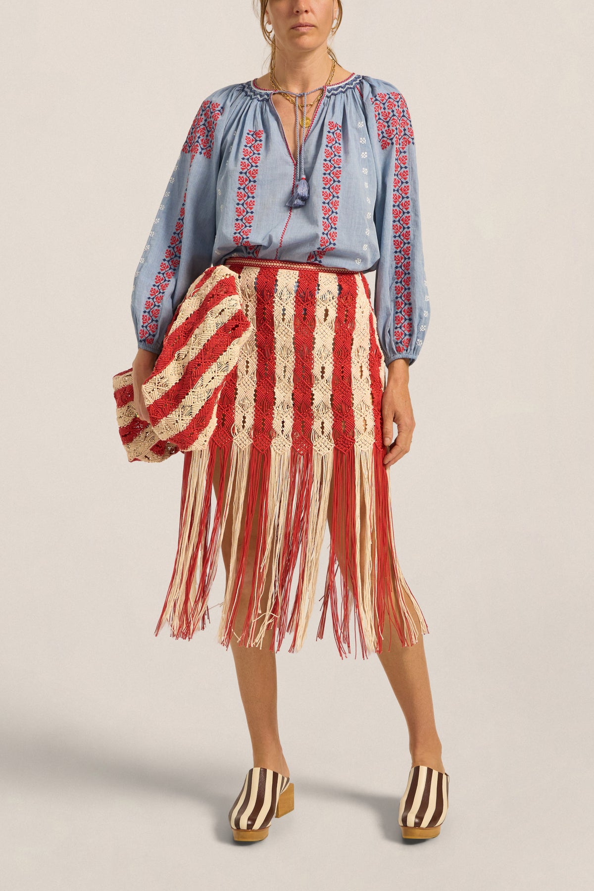 Person wearing a blue embroidered blouse and red and white striped skirt with fringe on a plain background