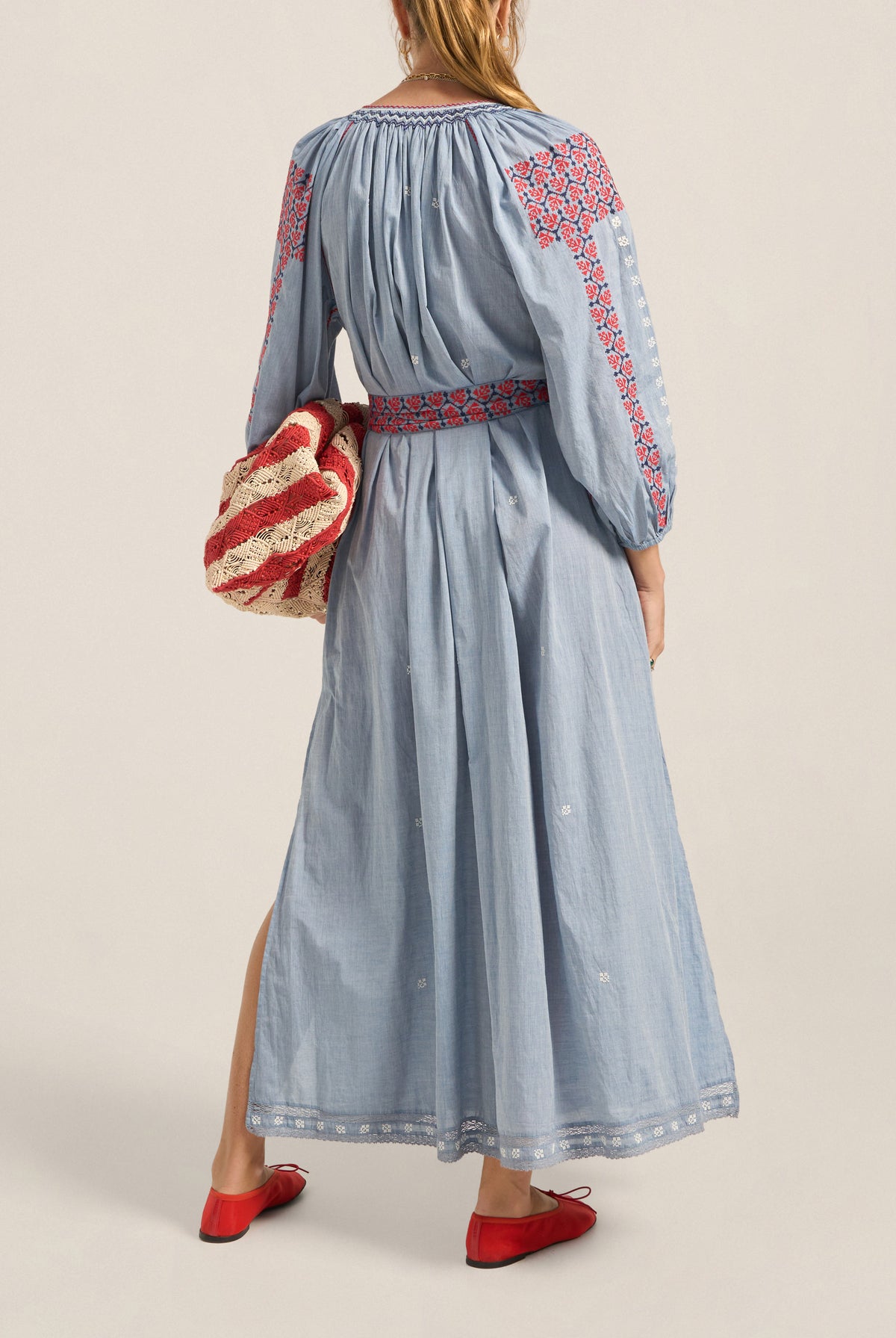 Woman wearing a long blue dress with red embroidery, holding a matching bag against a neutral background.