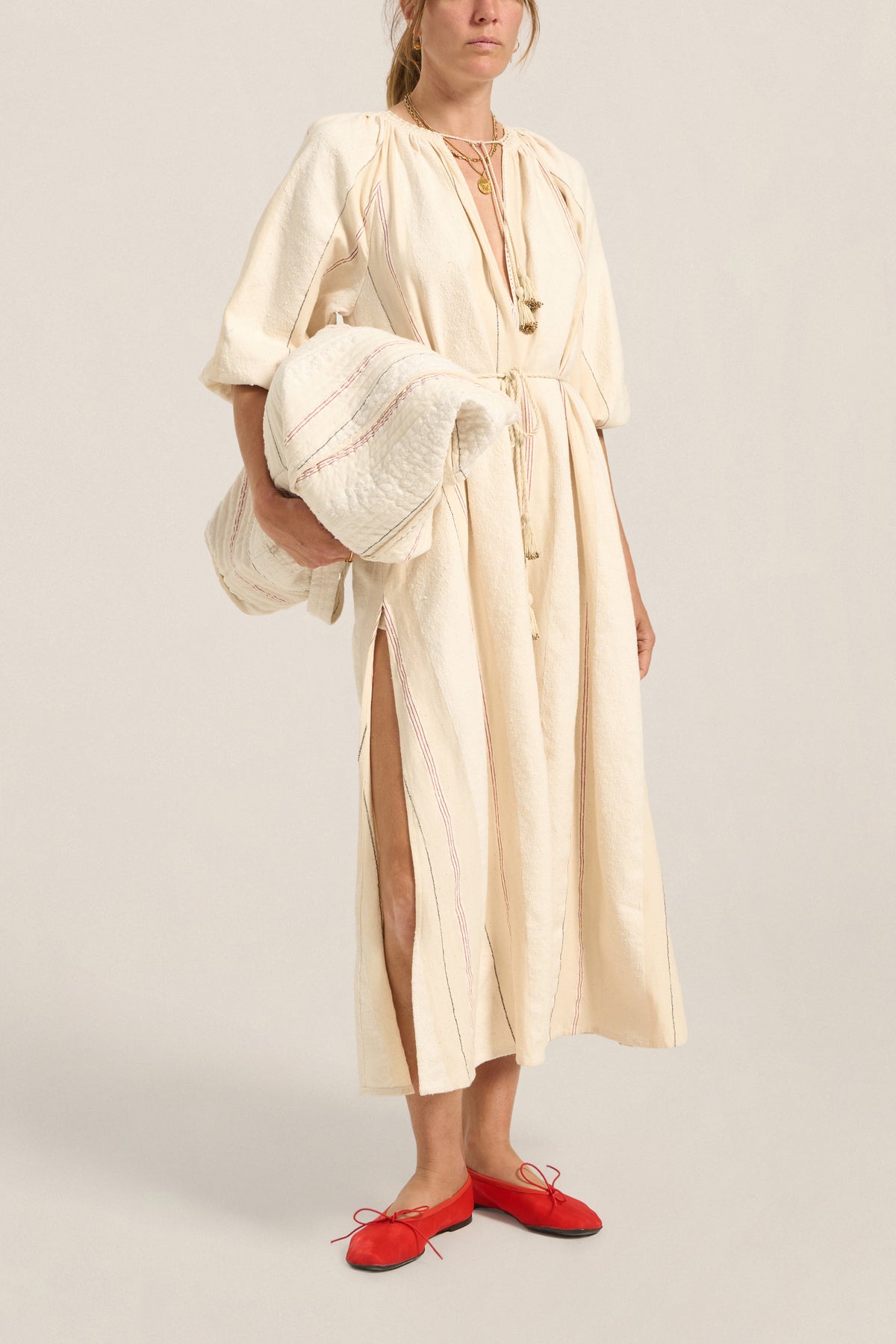 Woman wearing a long beige dress with a white hat and red shoes on a plain background