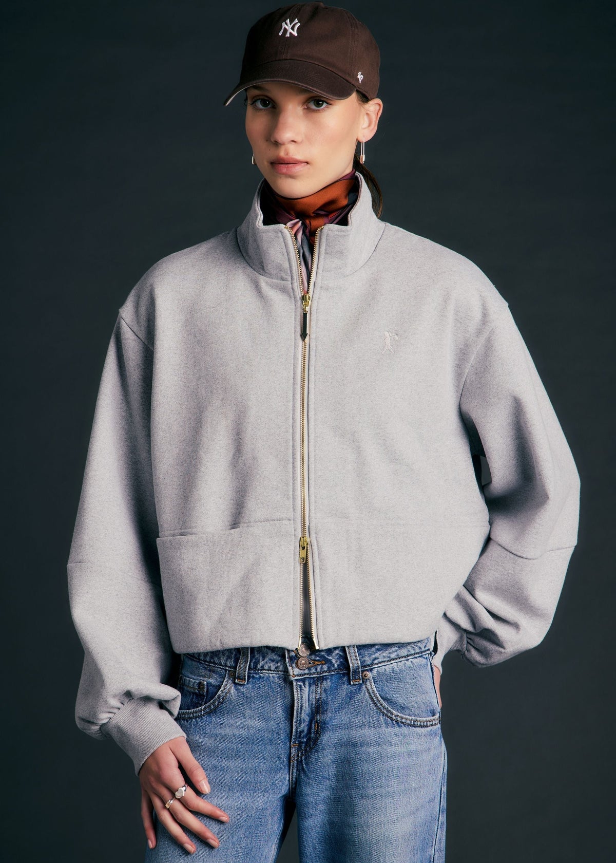 A model wearing a light gray cropped zip-up jacket, blue jeans, a brown New York Yankees baseball cap, and a rust-colored scarf, posed against a dark gray studio background.
