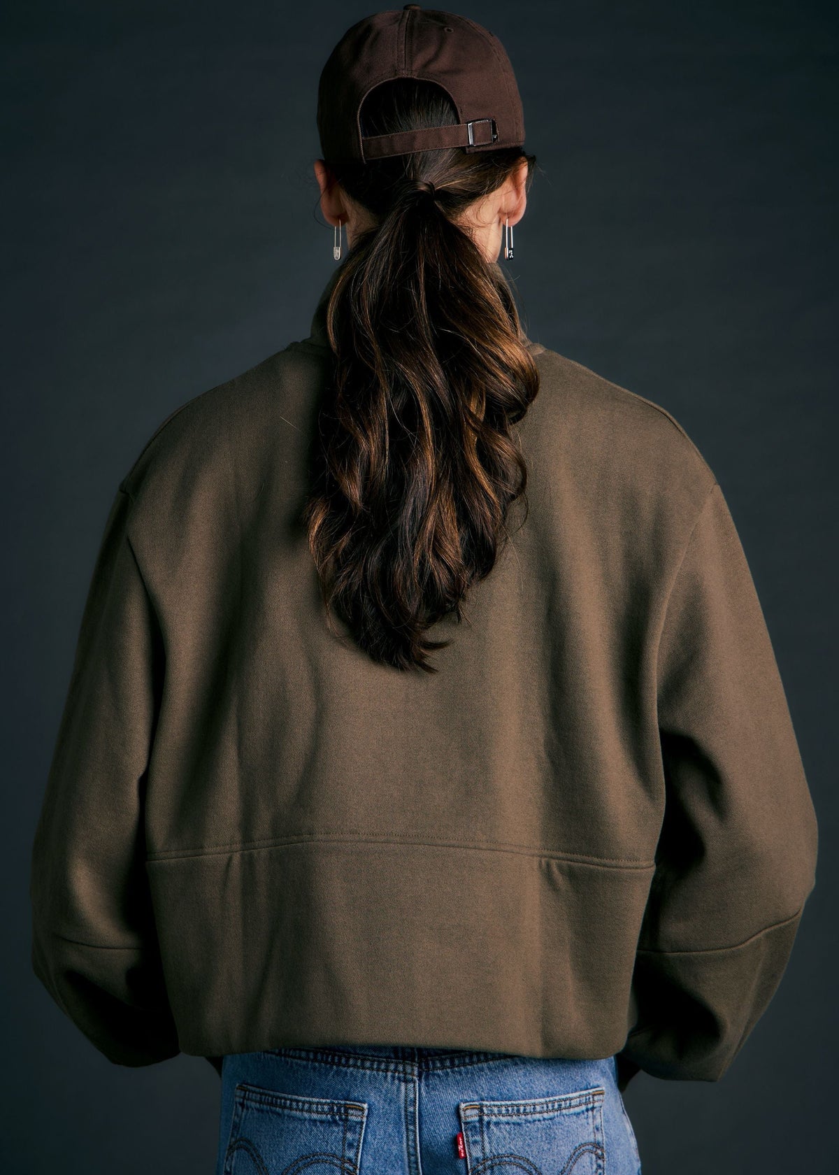 A person shown from the back wearing an oversized brown sweatshirt, a dark brown baseball cap, and blue jeans against a dark studio background.