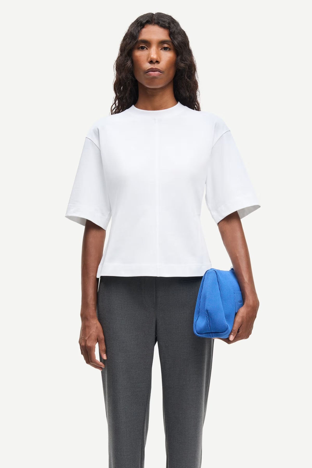 Woman wearing a white top and gray pants, holding a blue clutch against a white background