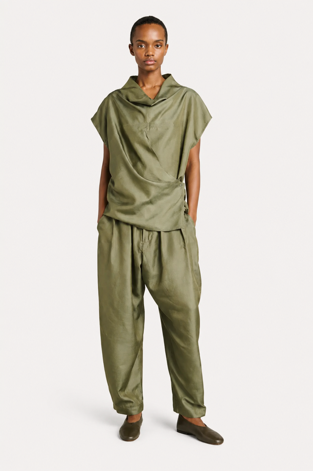A model wearing a matching olive green draped two-piece outfit consisting of a short-sleeve cowl-neck top and relaxed drawstring pants, paired with dark brown loafers, against a plain light gray background.
