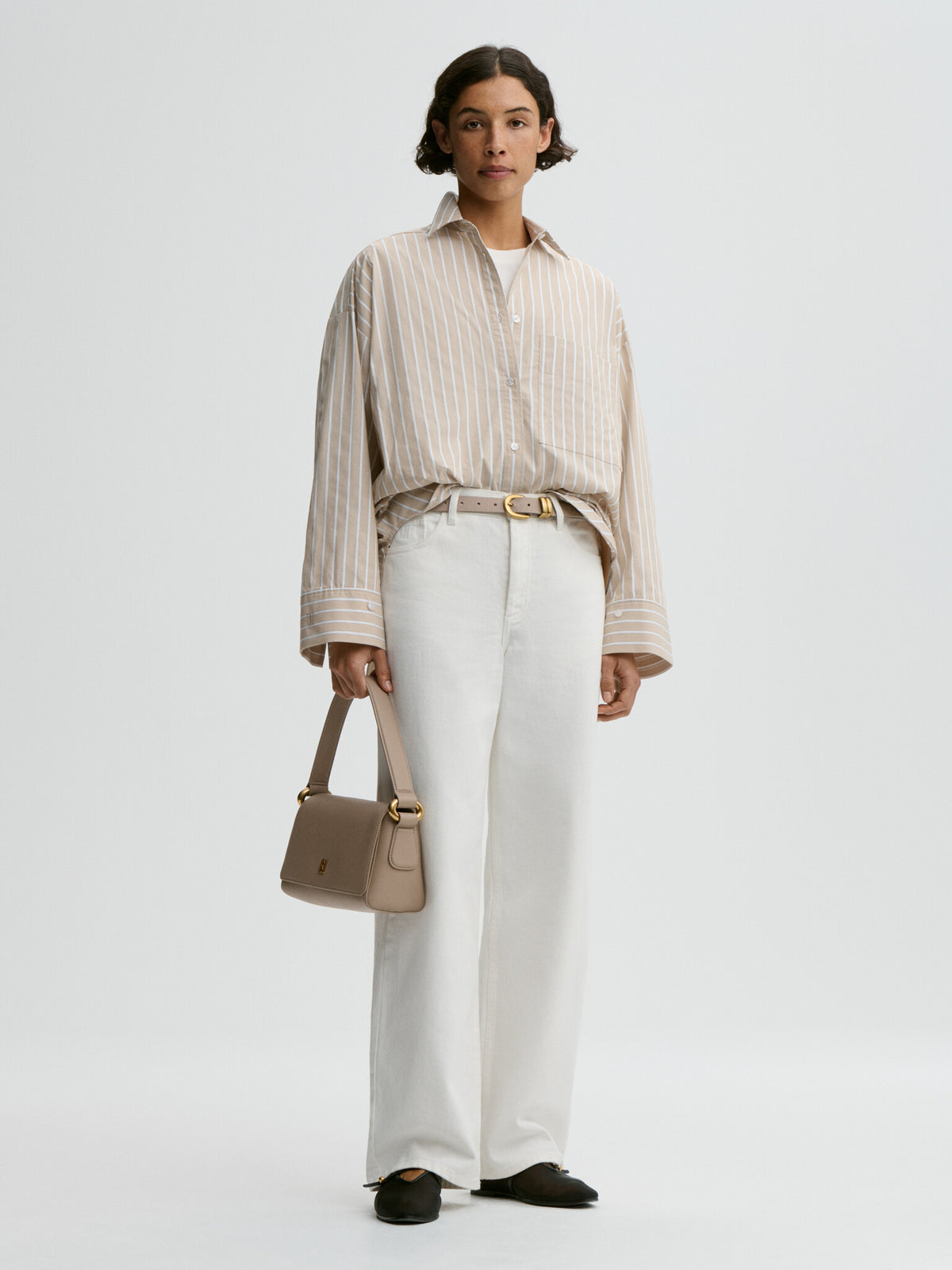 Woman wearing a beige striped shirt and white pants, holding a beige handbag on a light gray background