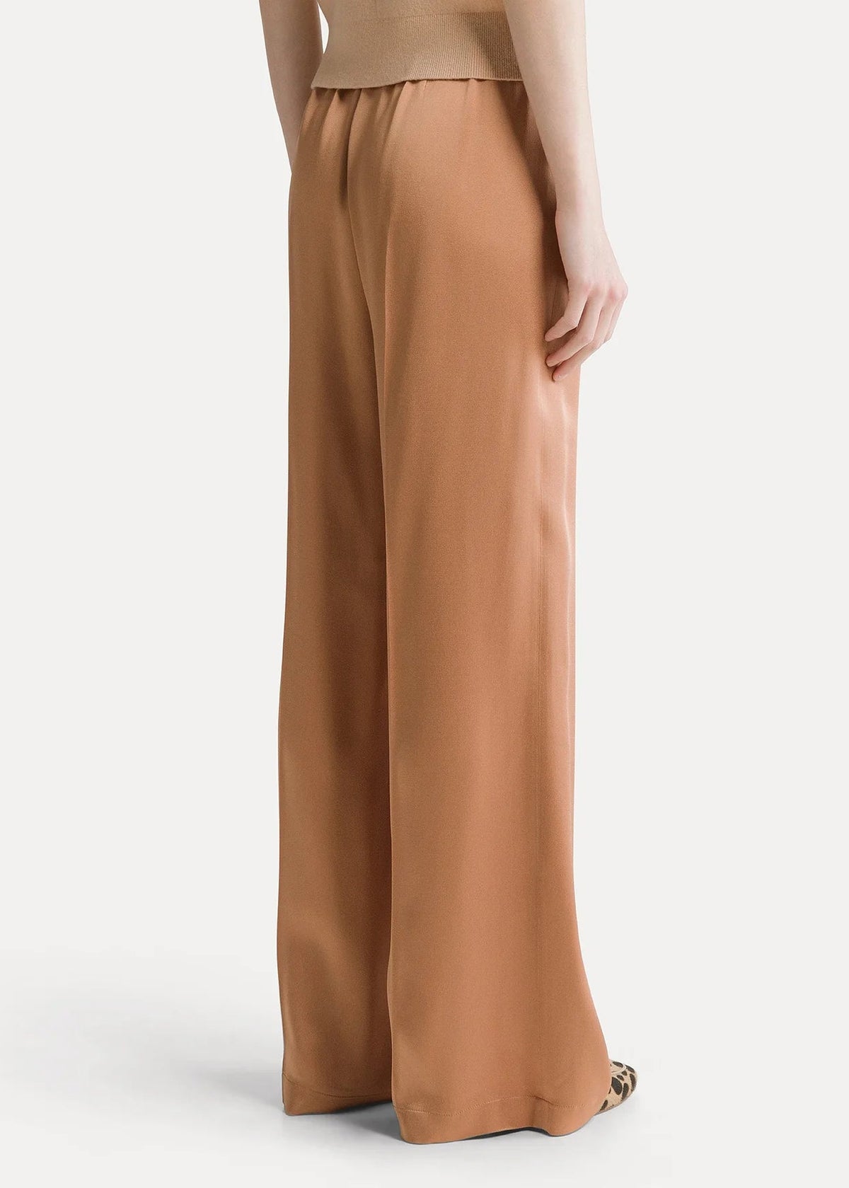 A model shown from the back wearing wide-leg trousers in a warm camel brown color, paired with a matching beige top, against a light gray studio background.