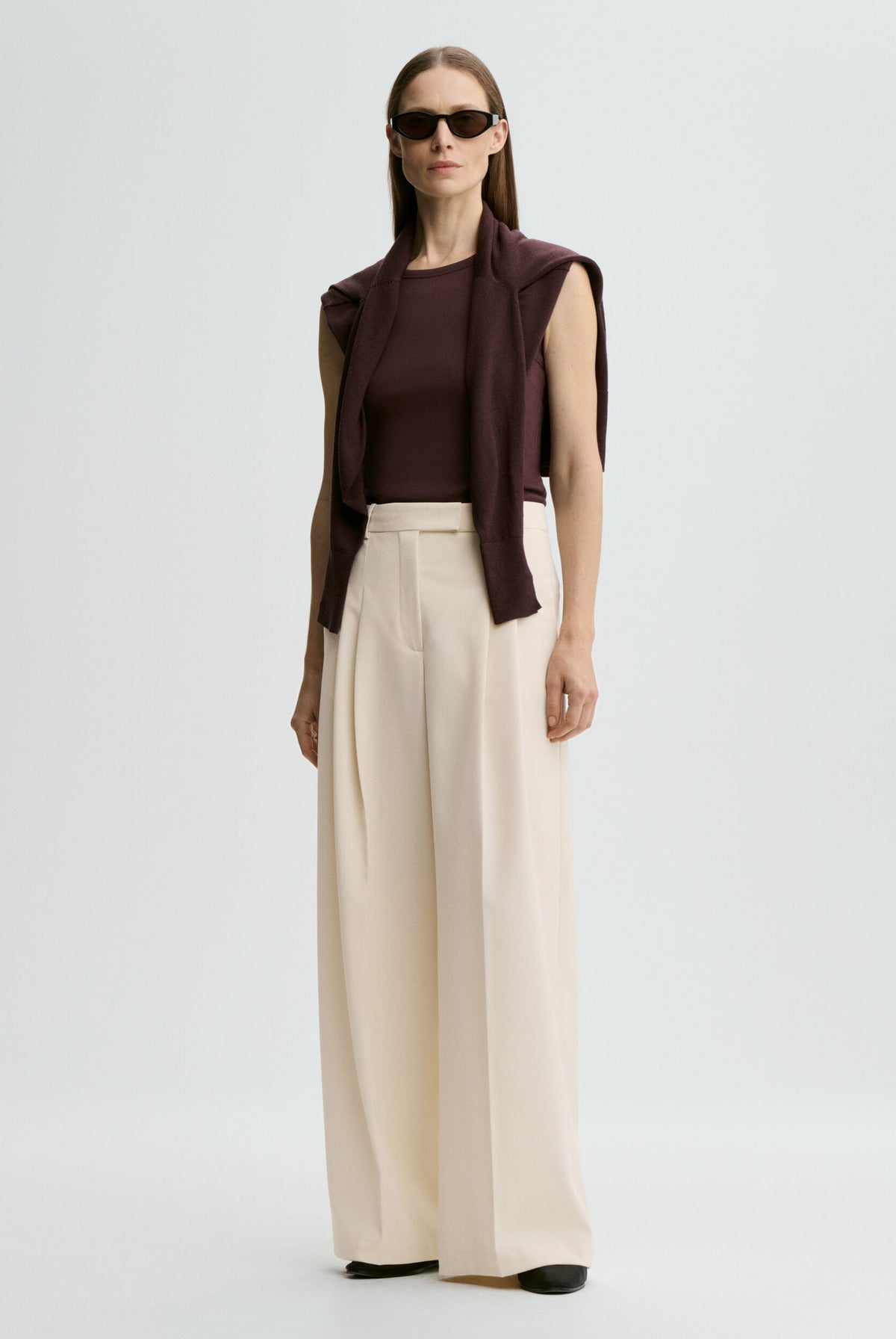 Woman wearing a brown top, beige wide-leg pants, and sunglasses on a light gray background