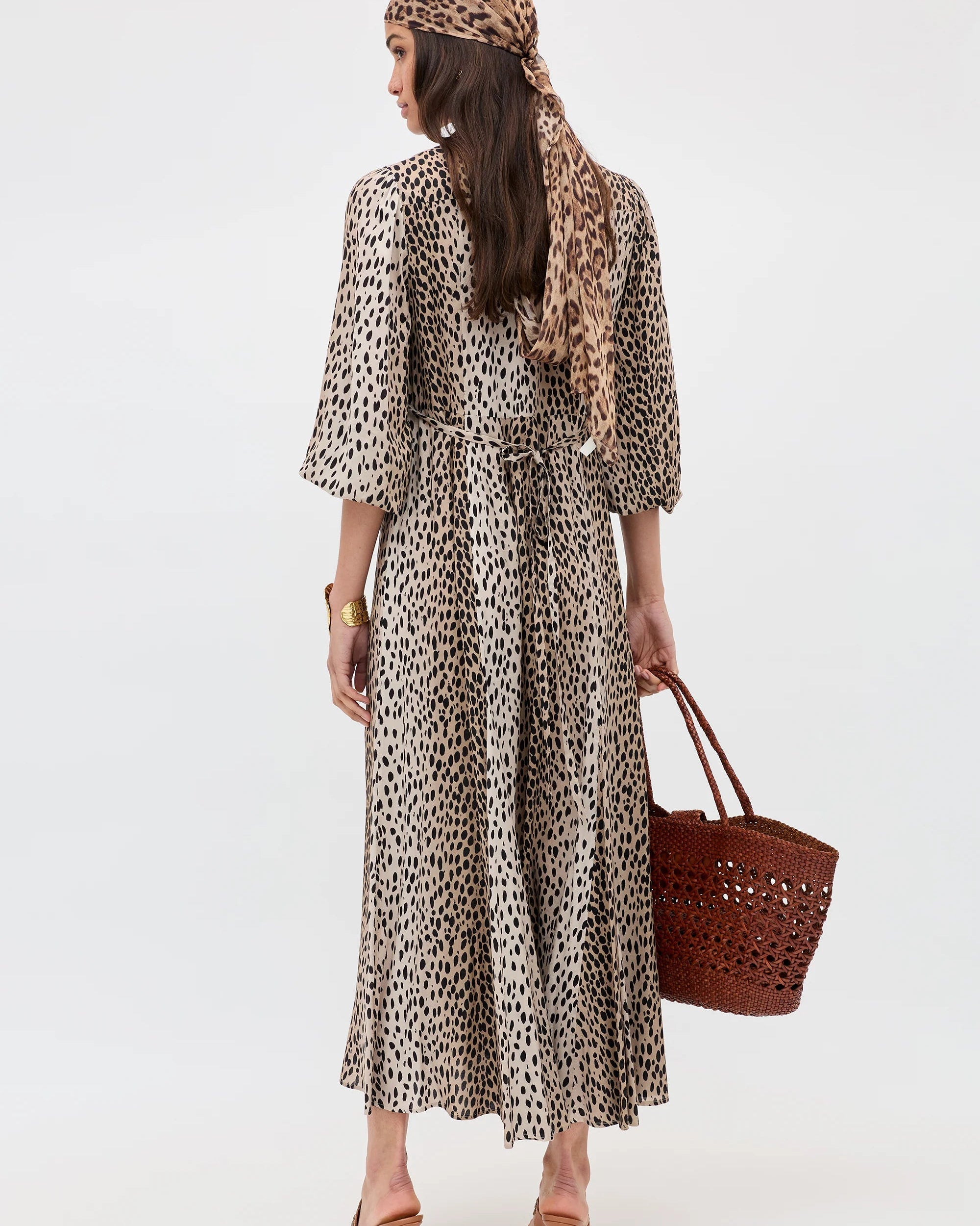 Woman wearing a leopard print dress and headscarf, holding a brown bag on a white background
