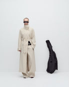 Person wearing a long beige coat with a black bag on a white background