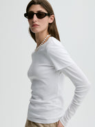 Woman wearing a white long-sleeve top and sunglasses on a light gray background