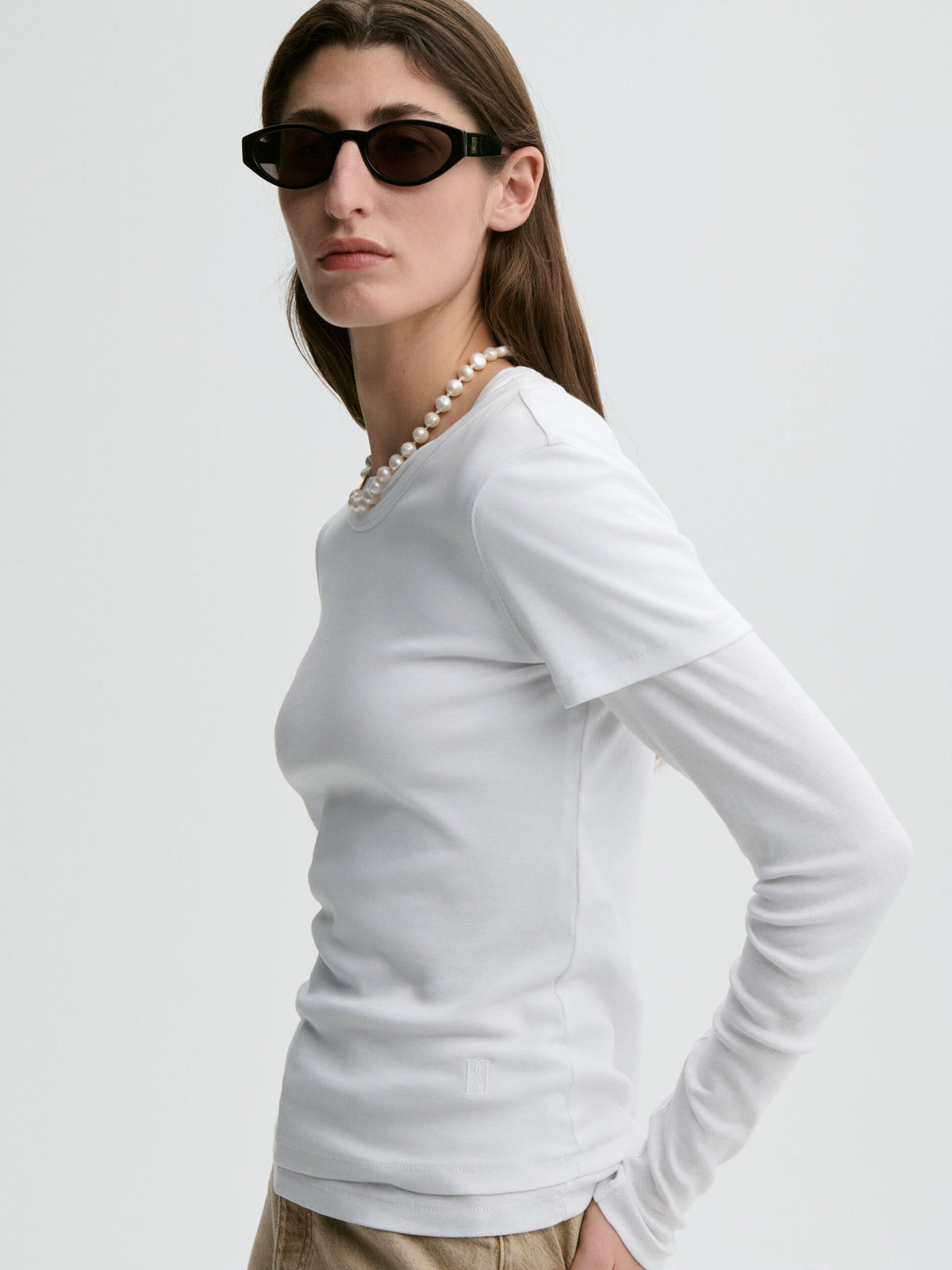 Woman wearing a white long-sleeve top and sunglasses on a light gray background