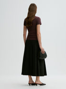 Woman wearing a brown top and black skirt holding a black clutch against a white background