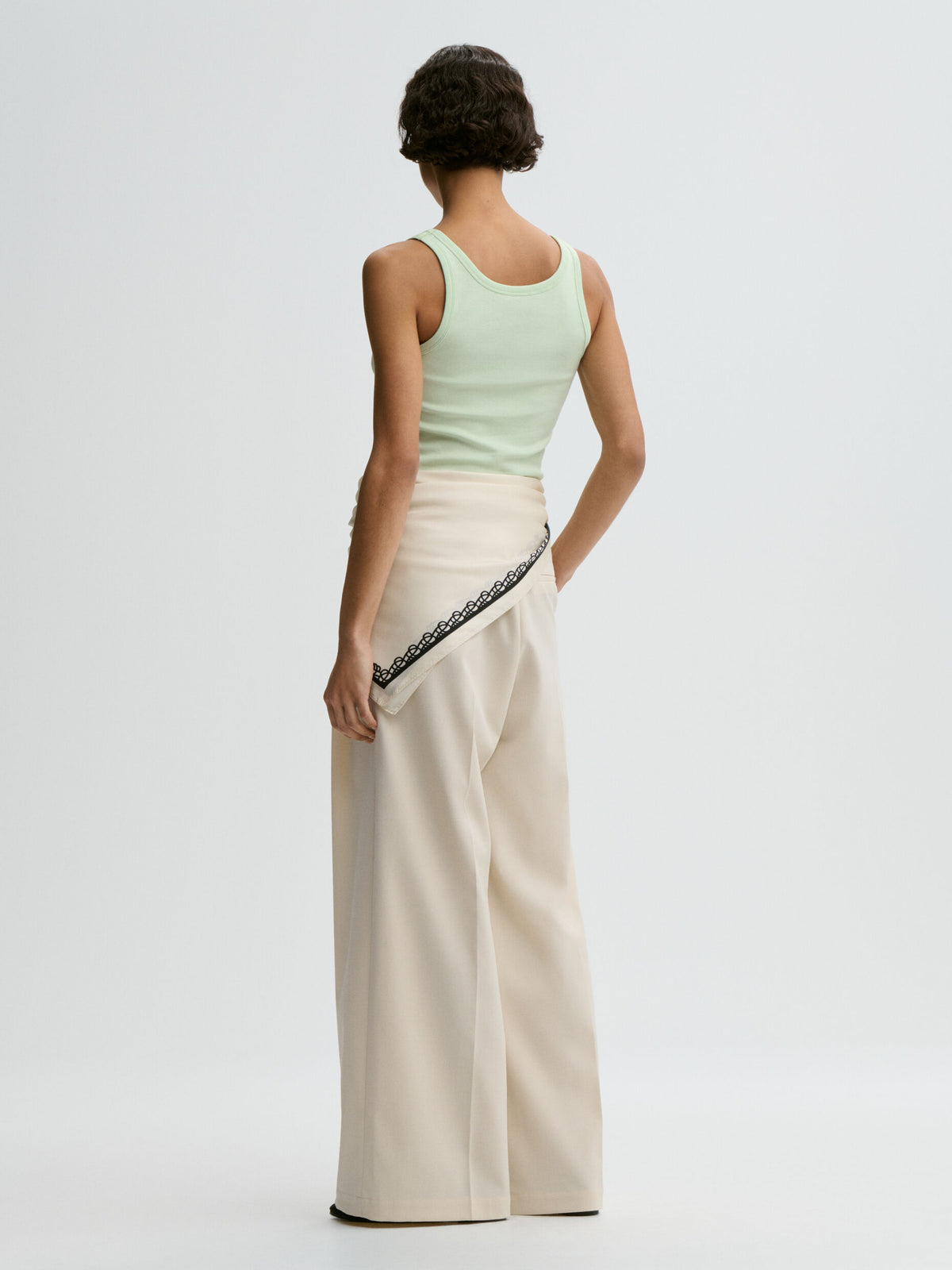 Woman wearing a light green sleeveless top and beige pants with lace trim on a white background
