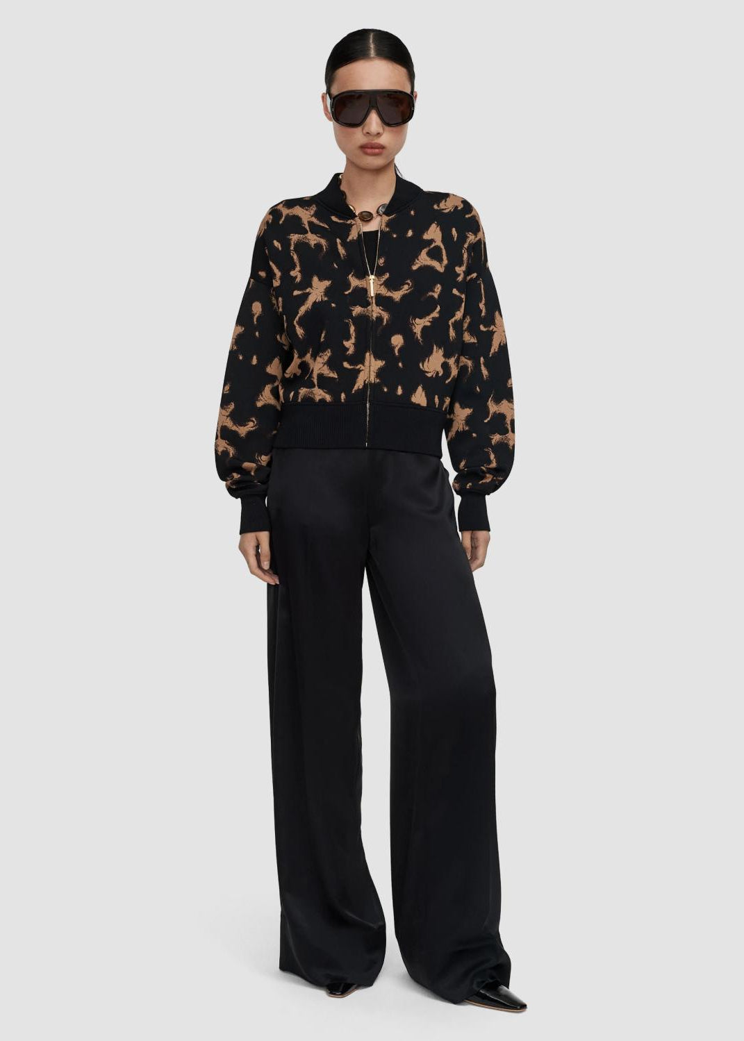 A model wearing a black and tan patterned bomber jacket with black wide-leg trousers, black sunglasses, and black pointed shoes, photographed against a solid light gray background.