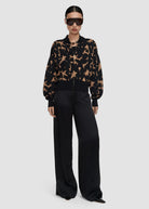 A model wearing a black and tan patterned bomber jacket with black wide-leg trousers, black sunglasses, and black pointed shoes, photographed against a solid light gray background.
