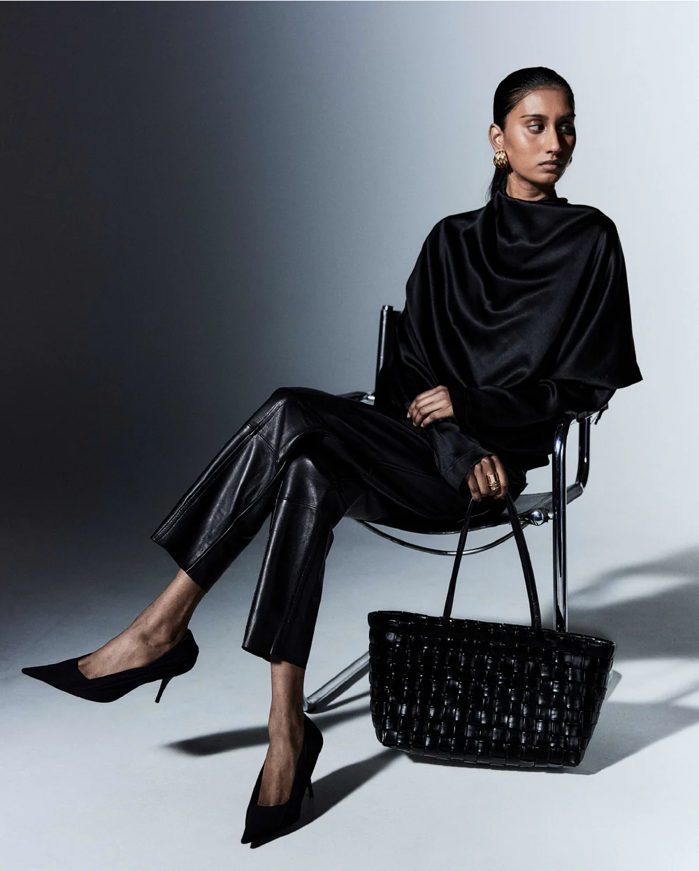 Woman in all-black outfit sitting on a chair with a black handbag beside her against a gray background