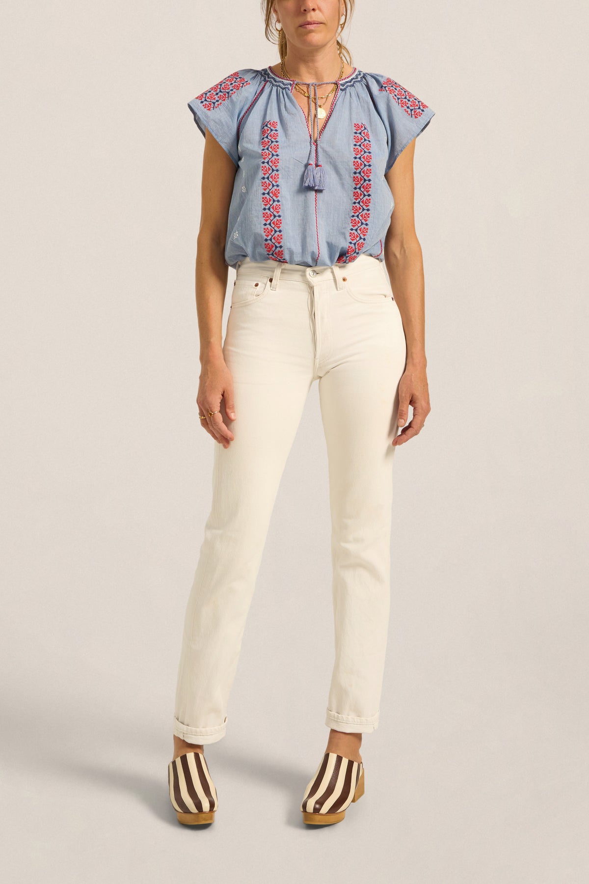 Woman wearing a blue embroidered top and white pants on a beige background