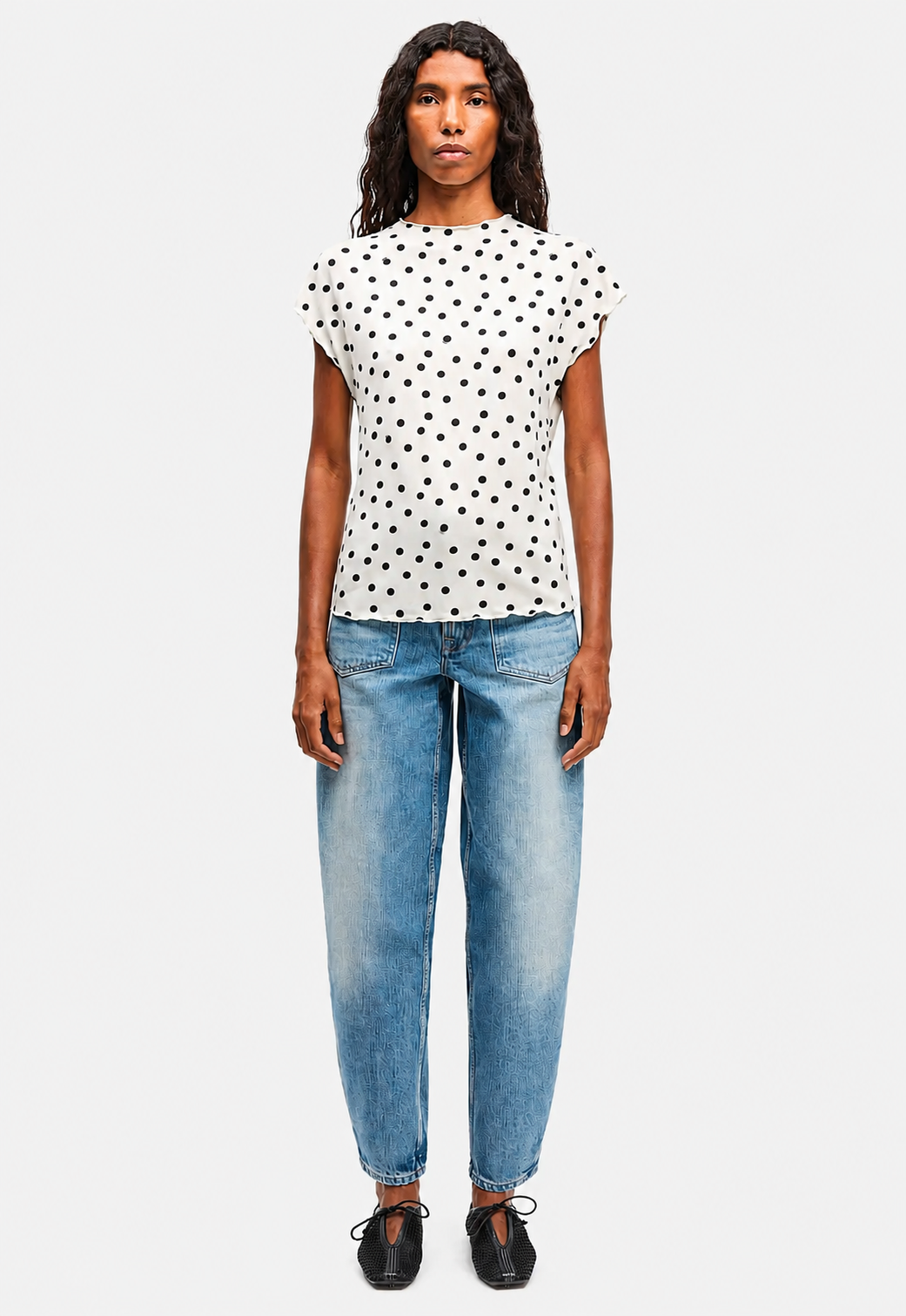 A woman modeling a white short-sleeve top with black polka dots, paired with light blue jeans and black shoes, against a light gray background.