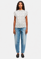 A woman modeling a white short-sleeve top with black polka dots, paired with light blue jeans and black shoes, against a light gray background.