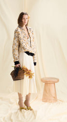 Woman in a floral jacket and white skirt holding flowers against a plain background
