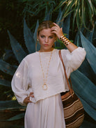Woman in a white outfit with gold jewelry and a striped bag against a green plant background