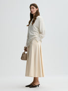 Woman wearing a white sweater and beige skirt holding a beige handbag on a light gray background