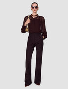 A model wearing a dark brown sheer long-sleeve blouse with a structured bodice and matching dark brown wide-leg trousers, styled with sunglasses, a chunky brown necklace, and a gold shoulder bag against a light gray background.