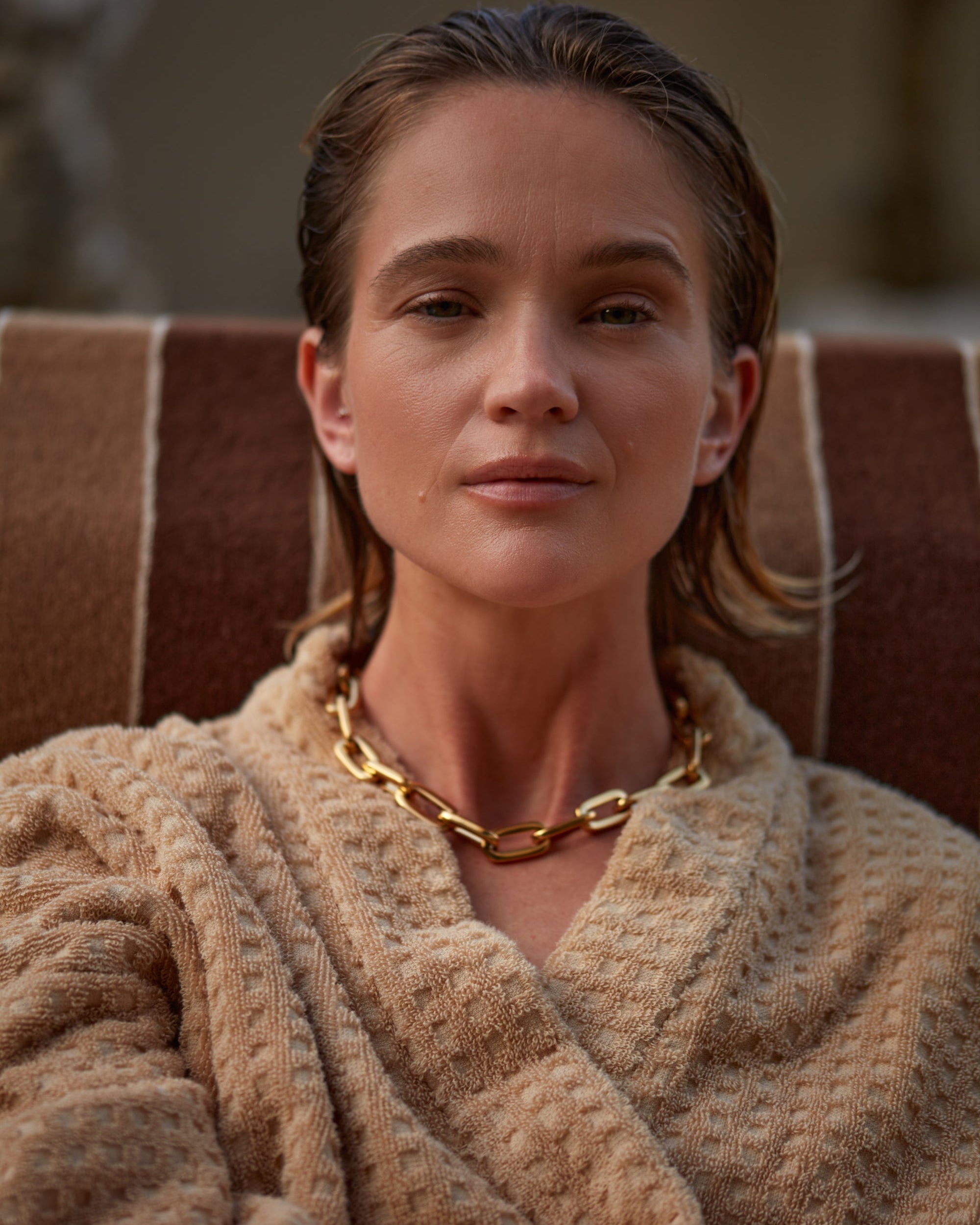 Woman wearing a beige robe and gold necklace, sitting on a brown chair.