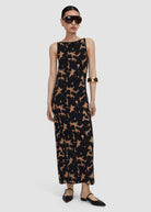 A model wearing a sleeveless black maxi dress with a tan abstract animal-like print, styled with black flats, sunglasses, and a gold bangle against a light gray studio background.