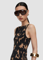 A model wearing a sleeveless black top with a tan abstract print, styled with oversized dark sunglasses, gold hoop earrings, and a wide gold cuff bracelet against a light gray background.