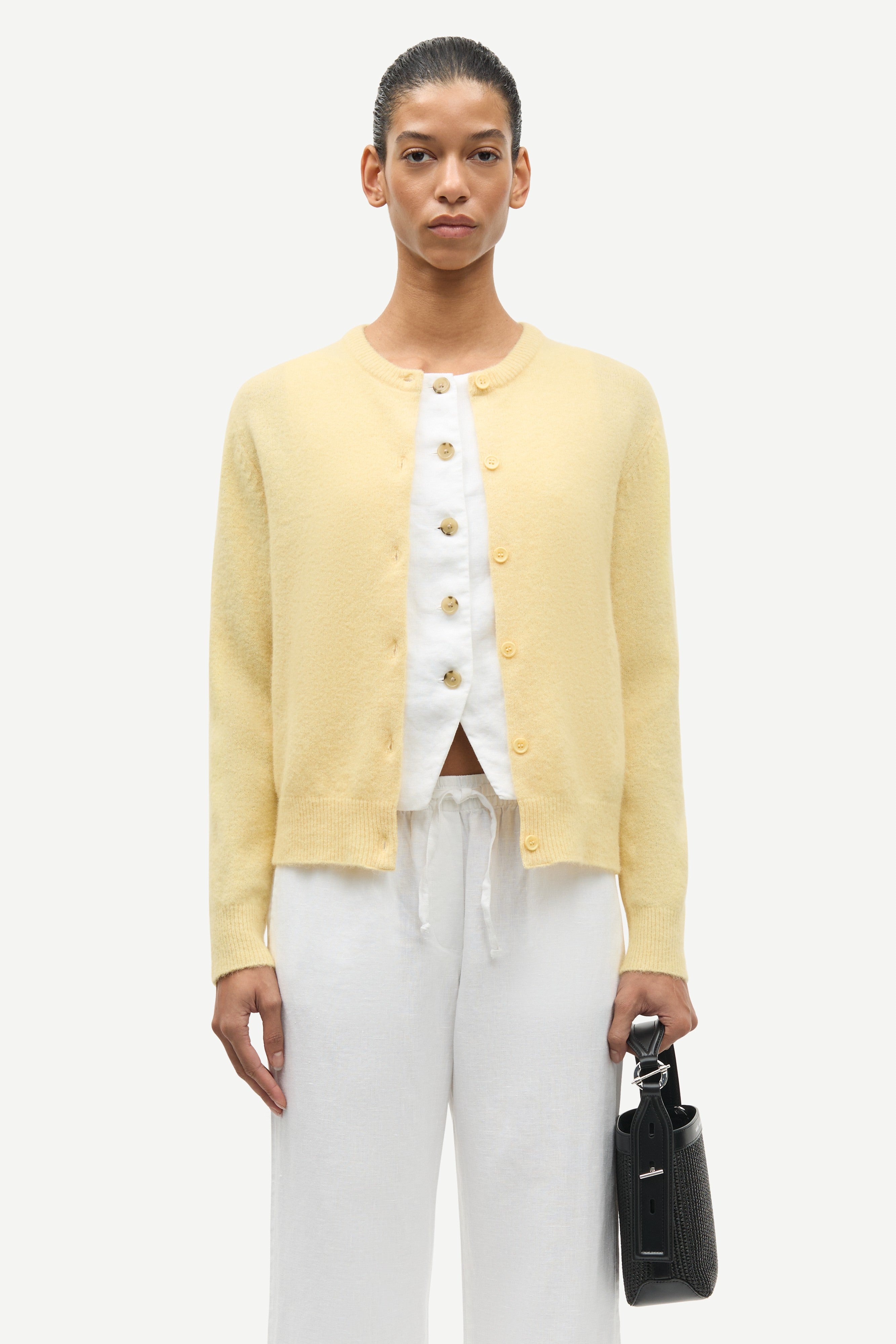 Person wearing a yellow cardigan over a white shirt with white pants, holding a black handbag on a white background.