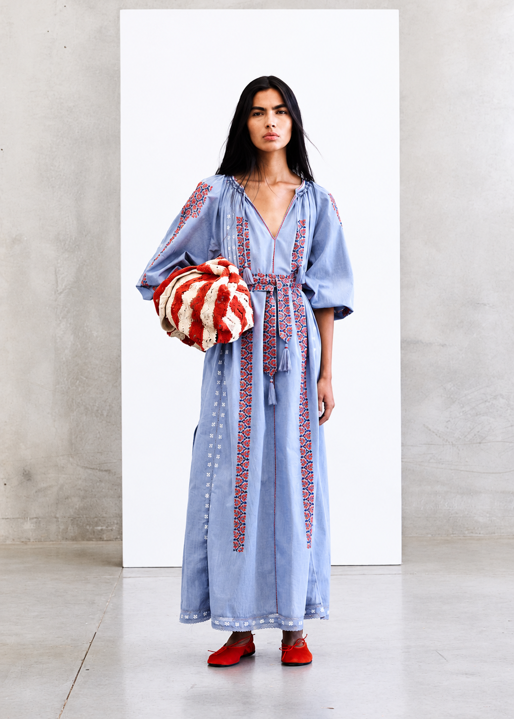 A model stands in a minimalist studio setting wearing a light blue maxi dress with red embroidered details and holding a red and white striped fuzzy bag.