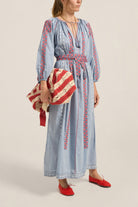 Woman wearing a blue dress with red embroidery, holding a red and white bag on a beige background