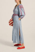 Woman wearing a light blue dress with red and white embroidery, holding a striped bag on a beige background.