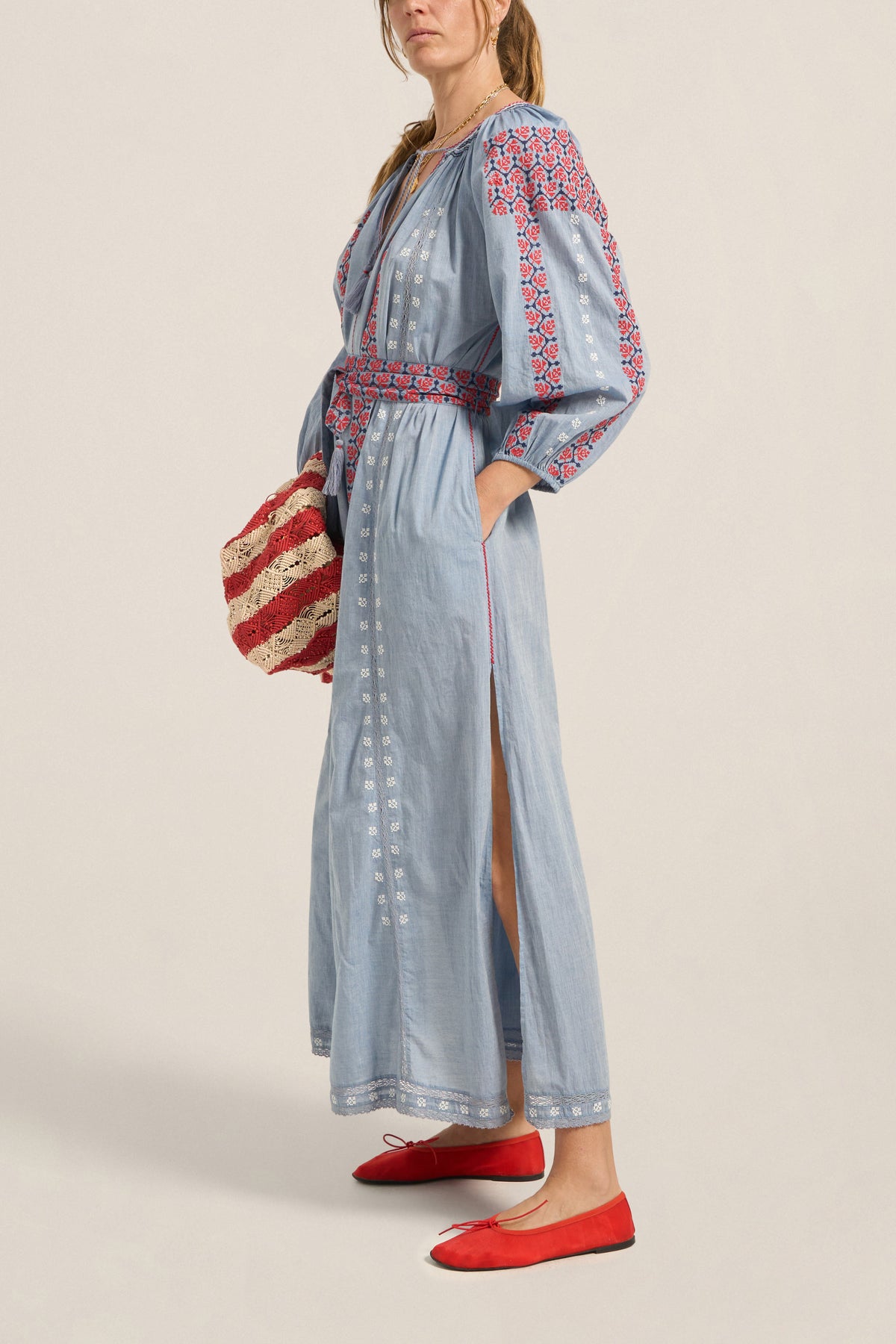 Woman wearing a light blue dress with red and white embroidery, holding a striped bag on a beige background.