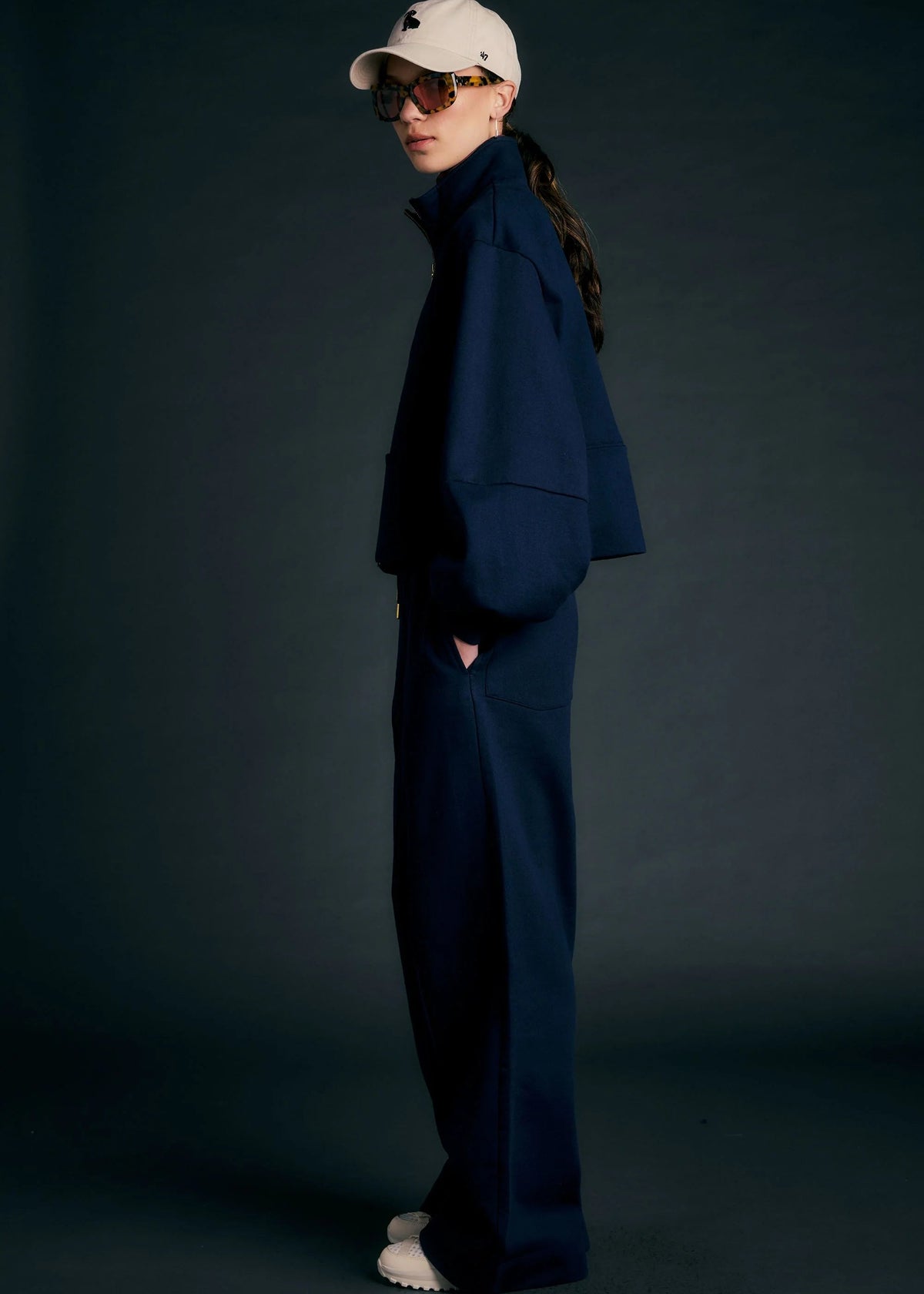 Person wearing a navy outfit with a white cap and sunglasses on a dark background