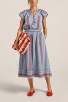 Woman wearing a blue dress with red and white embroidery, holding a red and white bag against a beige background.