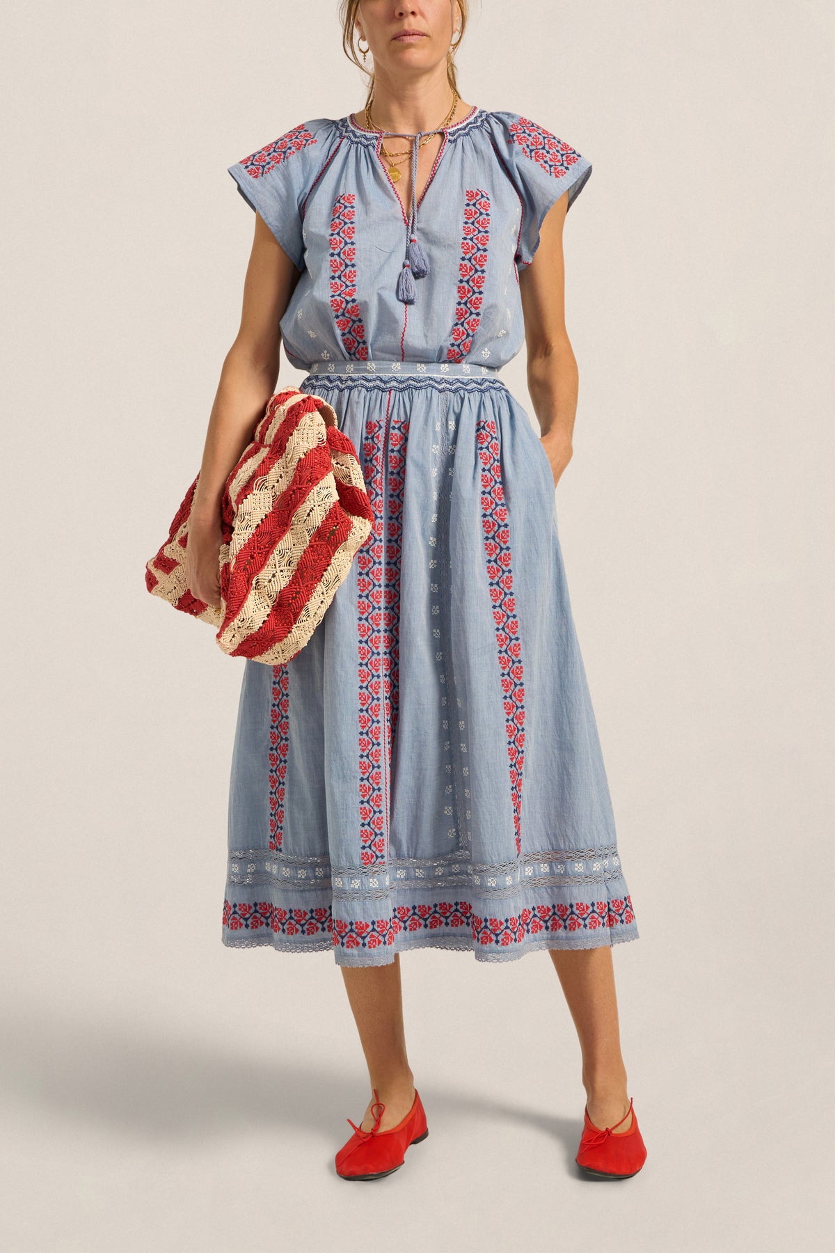 Woman wearing a blue dress with red and white embroidery, holding a red and white bag against a beige background.