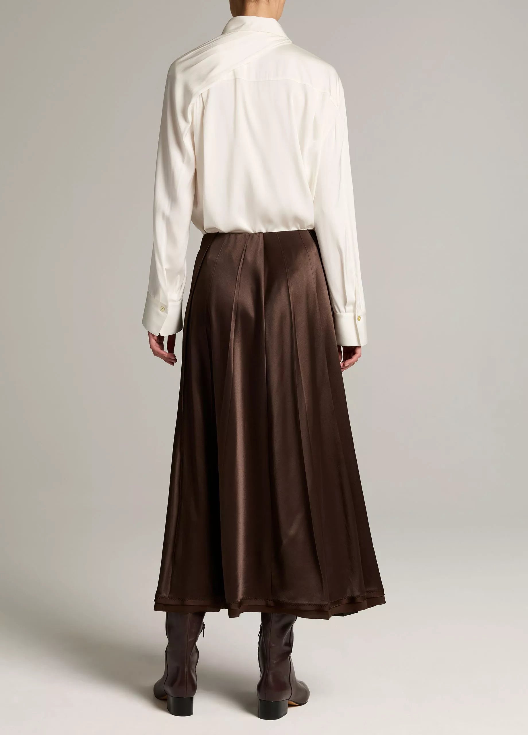 A model shown from the back wearing a white long-sleeve button-up shirt and a high-waisted, pleated brown satin midi skirt, paired with dark brown ankle boots, against a light gray studio background.