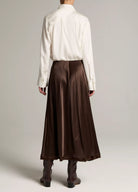 A model shown from the back wearing a white long-sleeve button-up shirt and a high-waisted, pleated brown satin midi skirt, paired with dark brown ankle boots, against a light gray studio background.