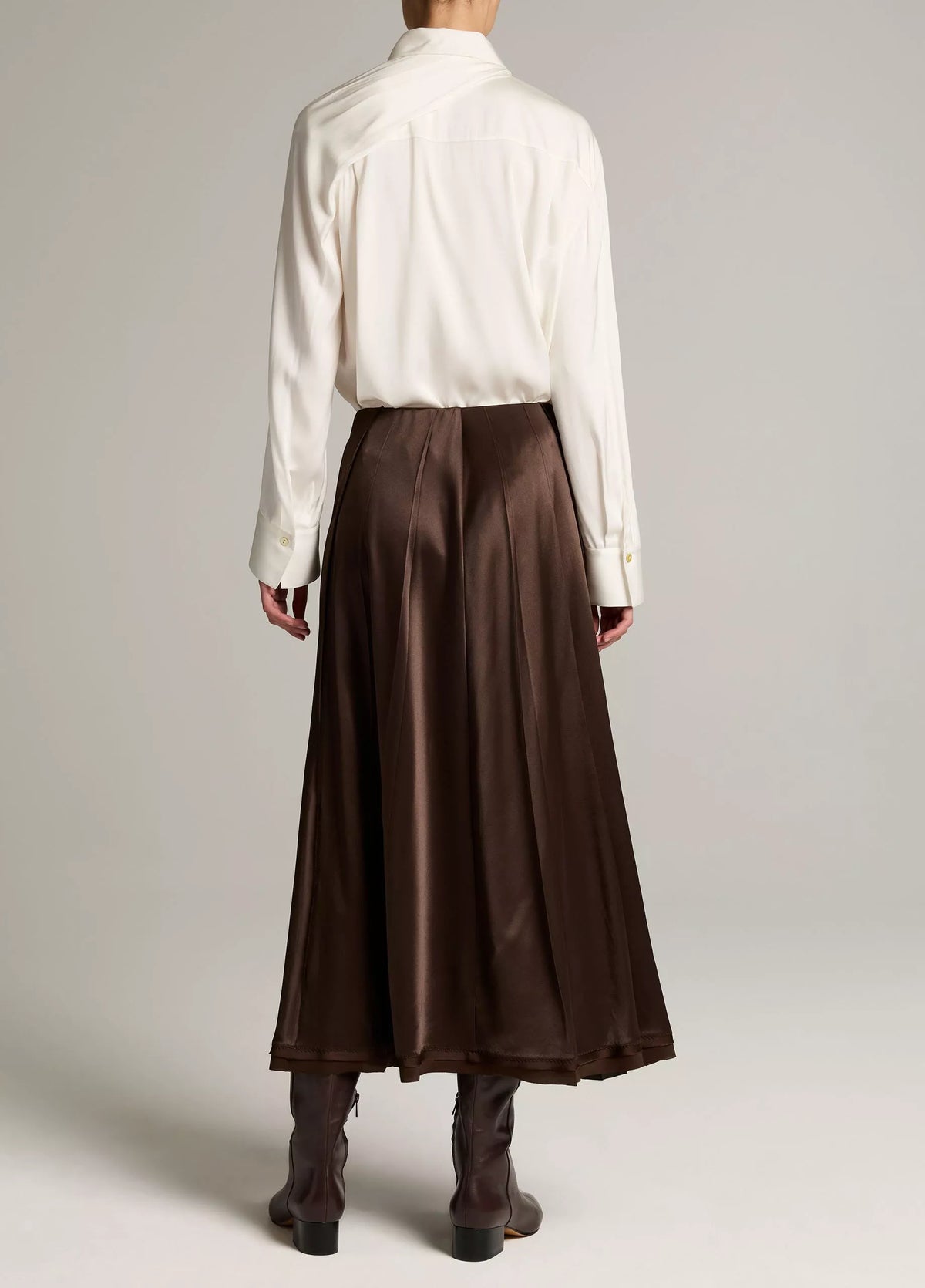 A model shown from the back wearing a white long-sleeve button-up shirt and a high-waisted, pleated brown satin midi skirt, paired with dark brown ankle boots, against a light gray studio background.