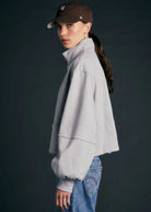 A woman wearing a light gray cropped quarter-zip sweatshirt, blue jeans, and a dark brown baseball cap, shown from a side profile against a dark gray studio background.
