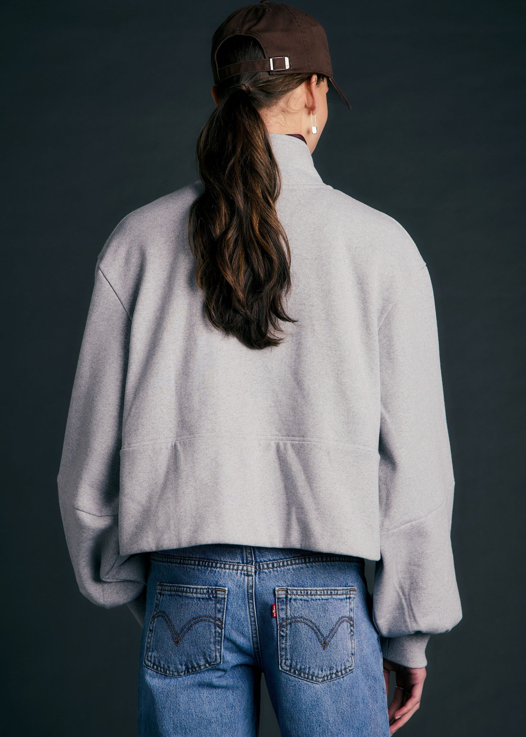 A person shown from the back wearing a light gray cropped quarter-zip fleece jacket, blue jeans, and a brown baseball cap against a dark gray studio background.