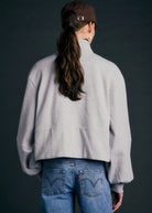 A person shown from the back wearing a light gray cropped quarter-zip fleece jacket, blue jeans, and a brown baseball cap against a dark gray studio background.