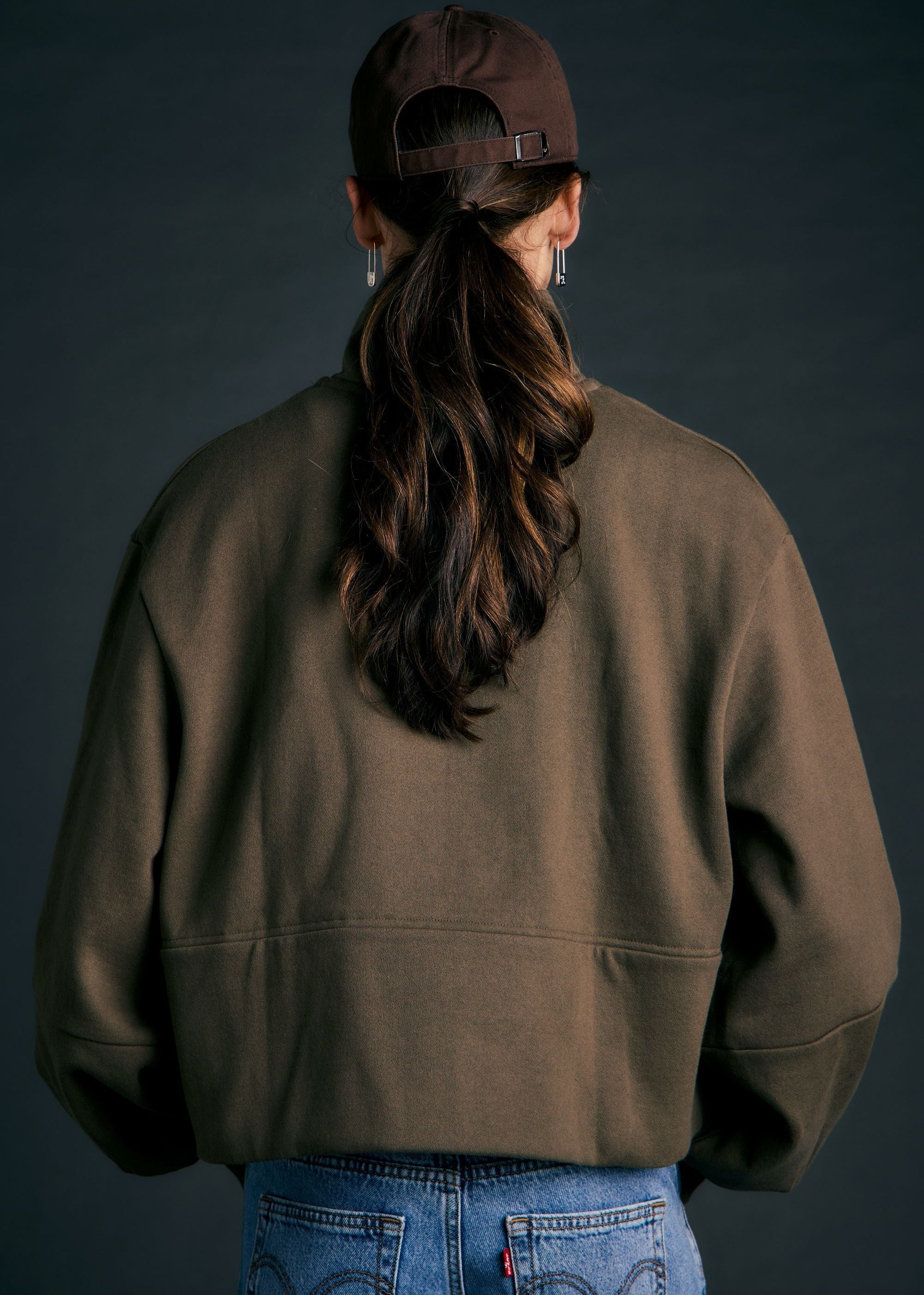A person shown from the back wearing an oversized brown sweatshirt, a dark brown baseball cap, and blue jeans against a dark studio background.