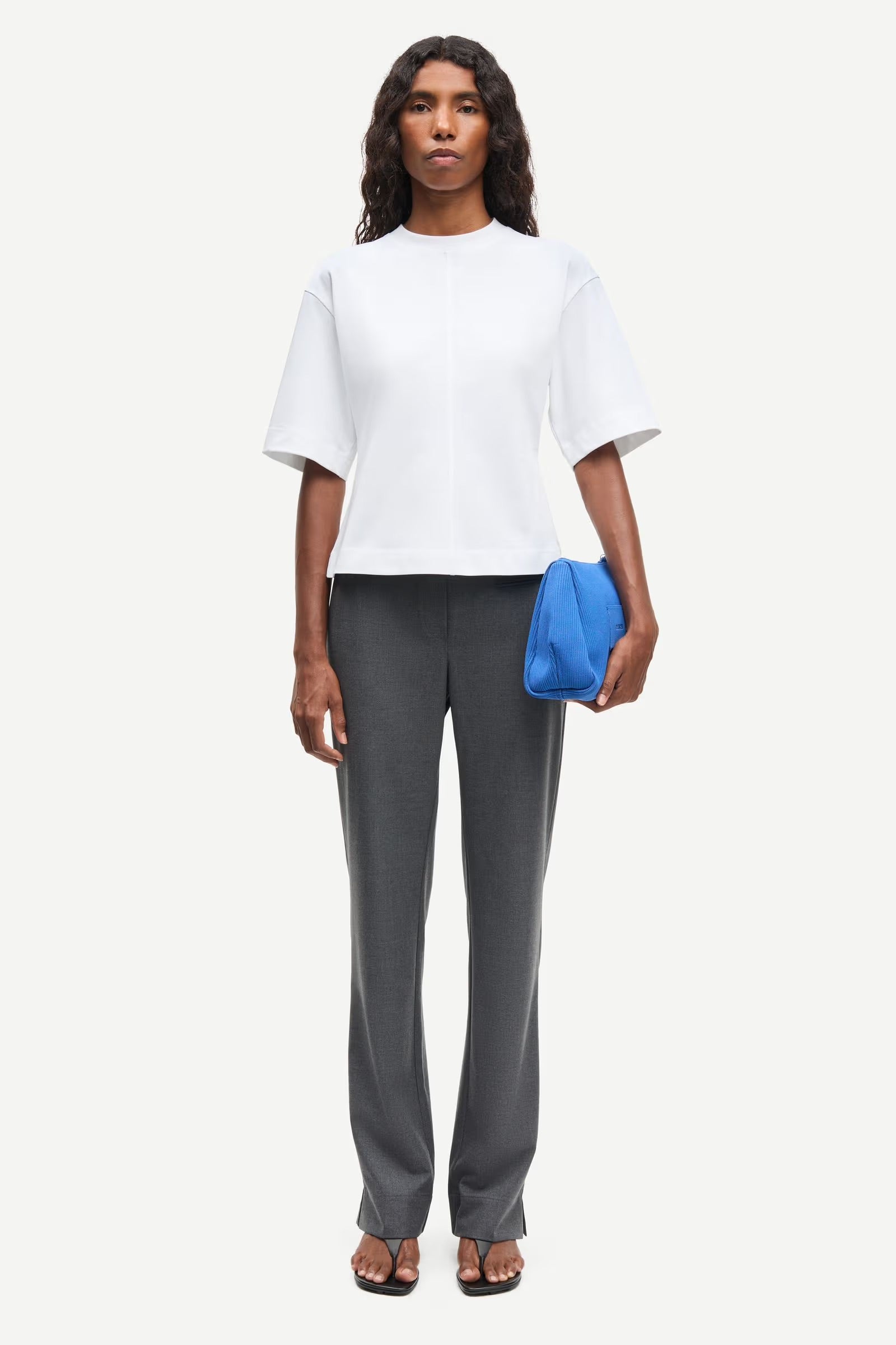 Woman wearing a white top and gray pants holding a blue clutch against a white background